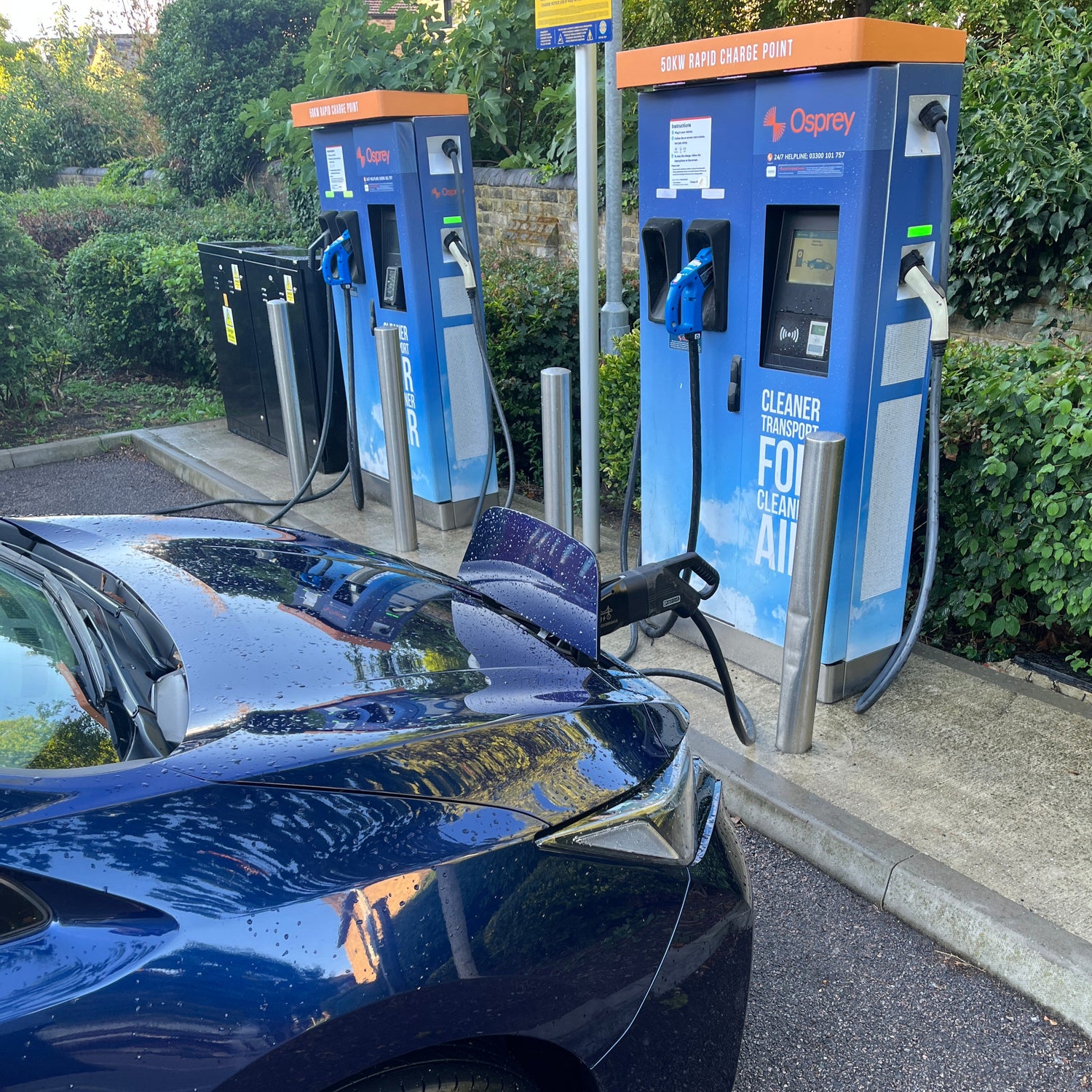Blue electric vehicle charging at a charging station with trees and pavement in the background